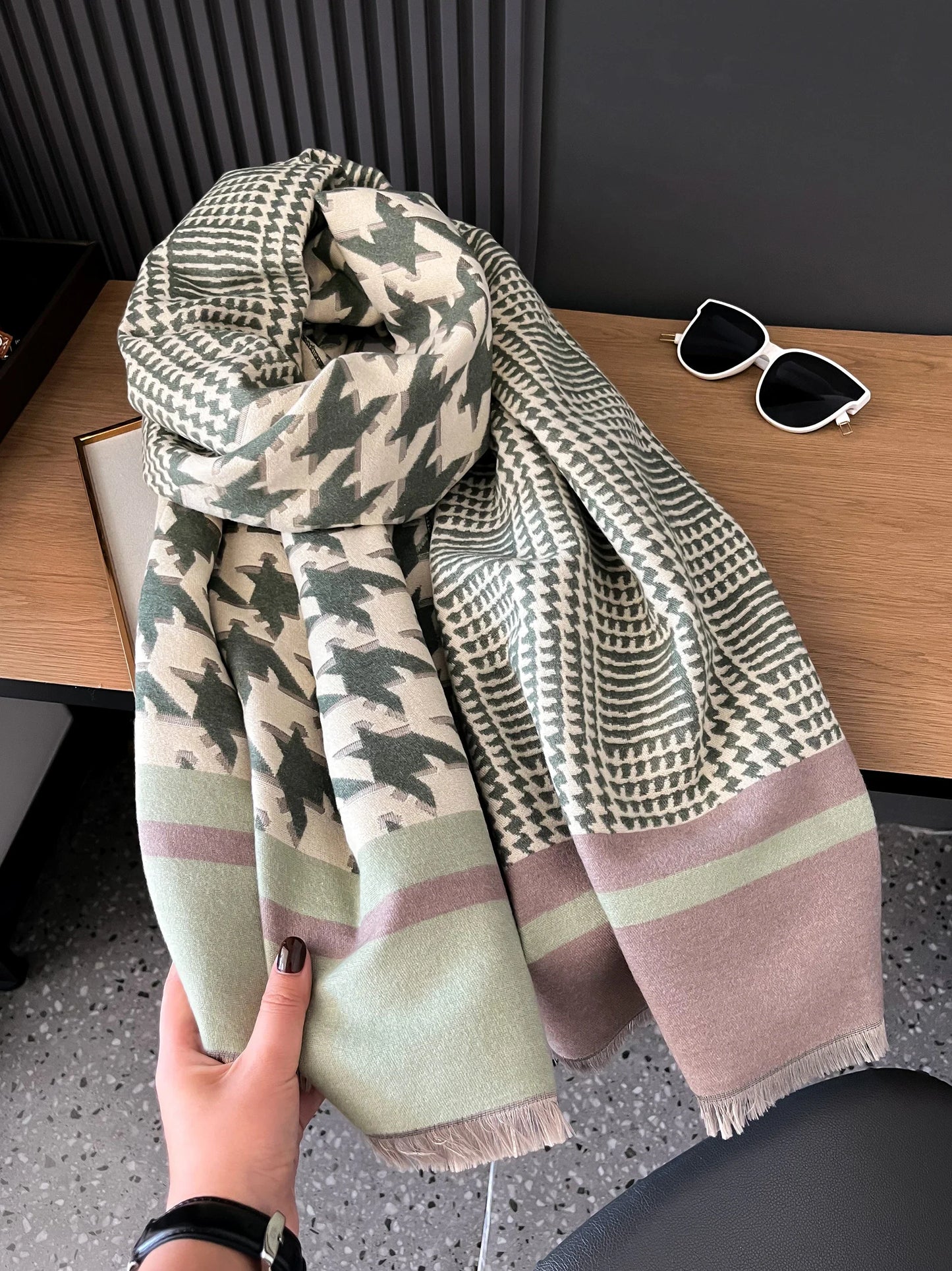 Women's cashmere scarf with houndstooth and geometric patterns in muted green and brown tones, styled on wooden surface with sunglasses.