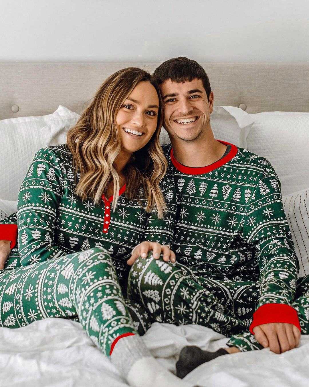 Couple wearing matching green and red Christmas family pajamas with holiday patterns sitting on bed smiling