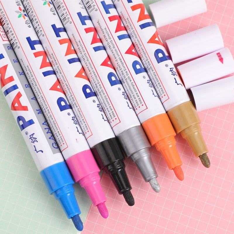 Set of vibrant paint markers with blue, pink, black, silver, orange, and gold tips on pink and green background