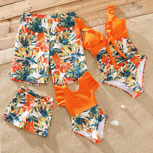 Family matching orange tropical plant print swimsuits and swim trunks set on sand background