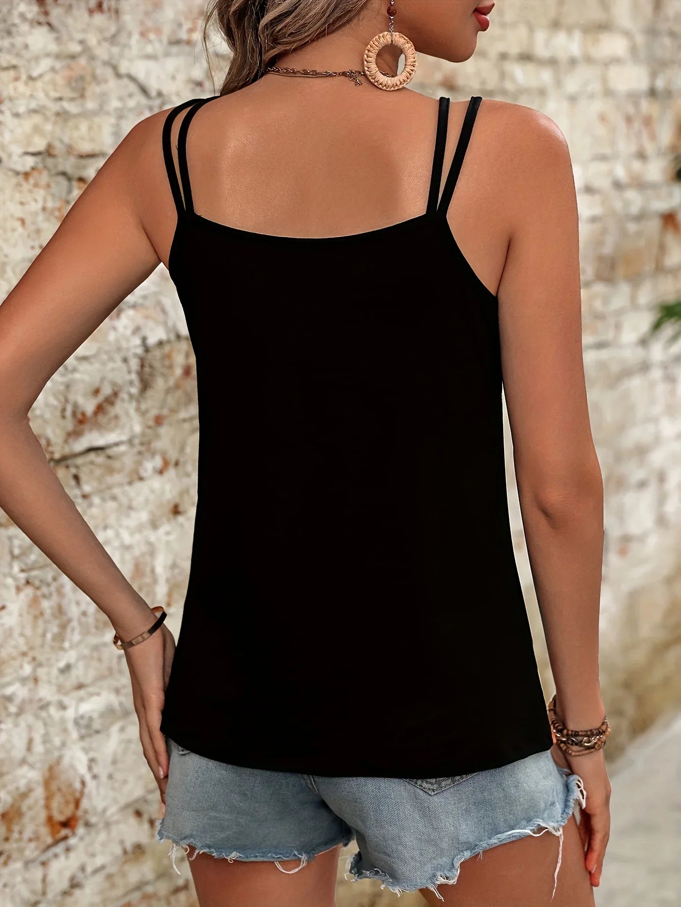 Back view of woman wearing black women's summer sleeveless tank top with double straps and denim shorts against brick wall