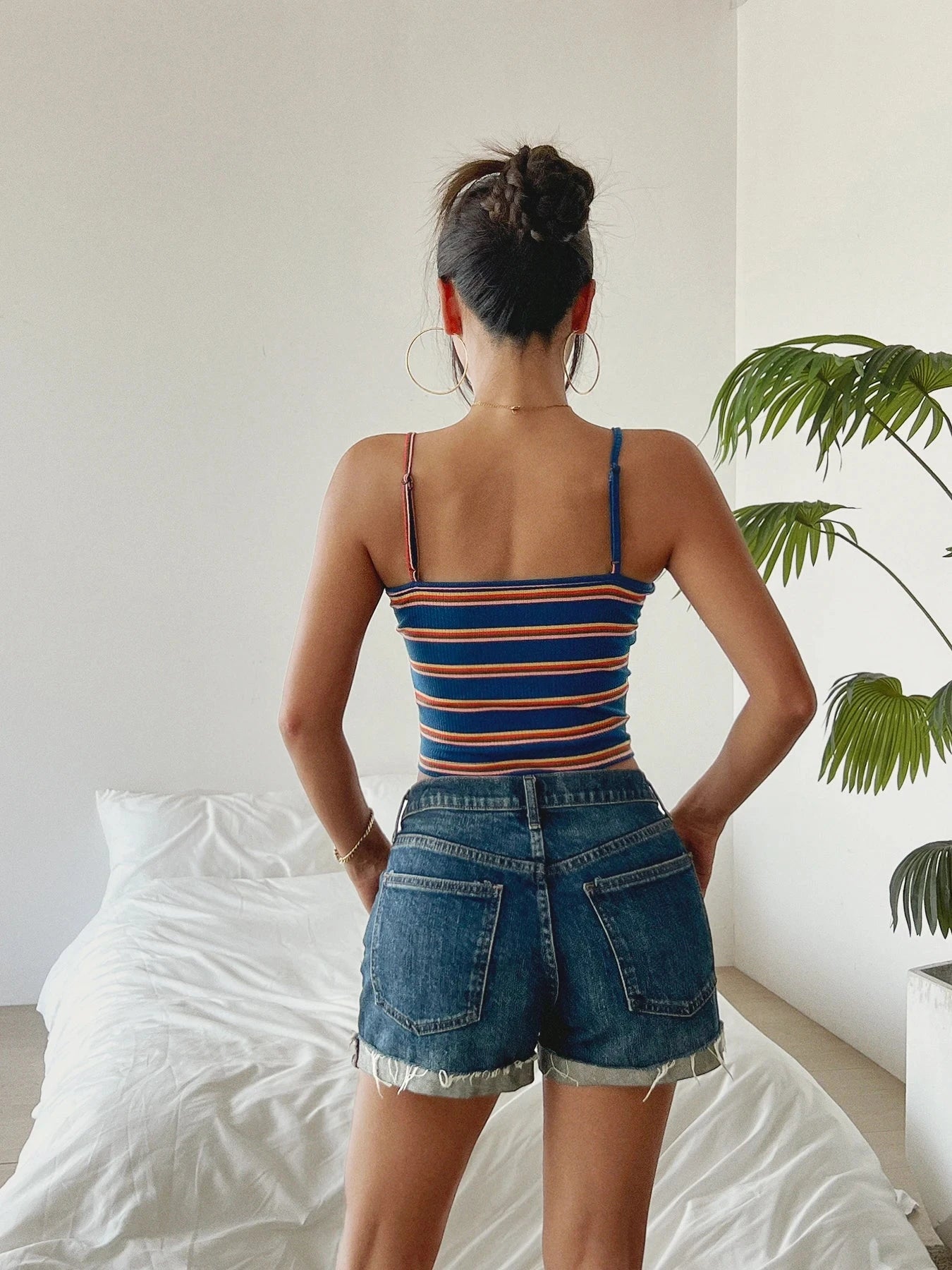 woman wearing striped women's summer tank top and denim shorts standing in bedroom with white bed and green plant