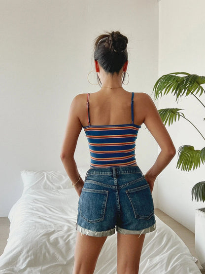 woman wearing striped women's summer tank top and denim shorts standing in bedroom with white bed and green plant